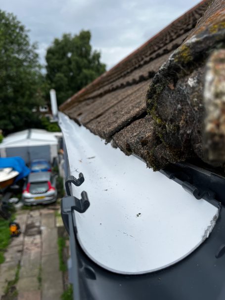 Close-up of a gutter along a roof edge, with trees and vehicles in the background.